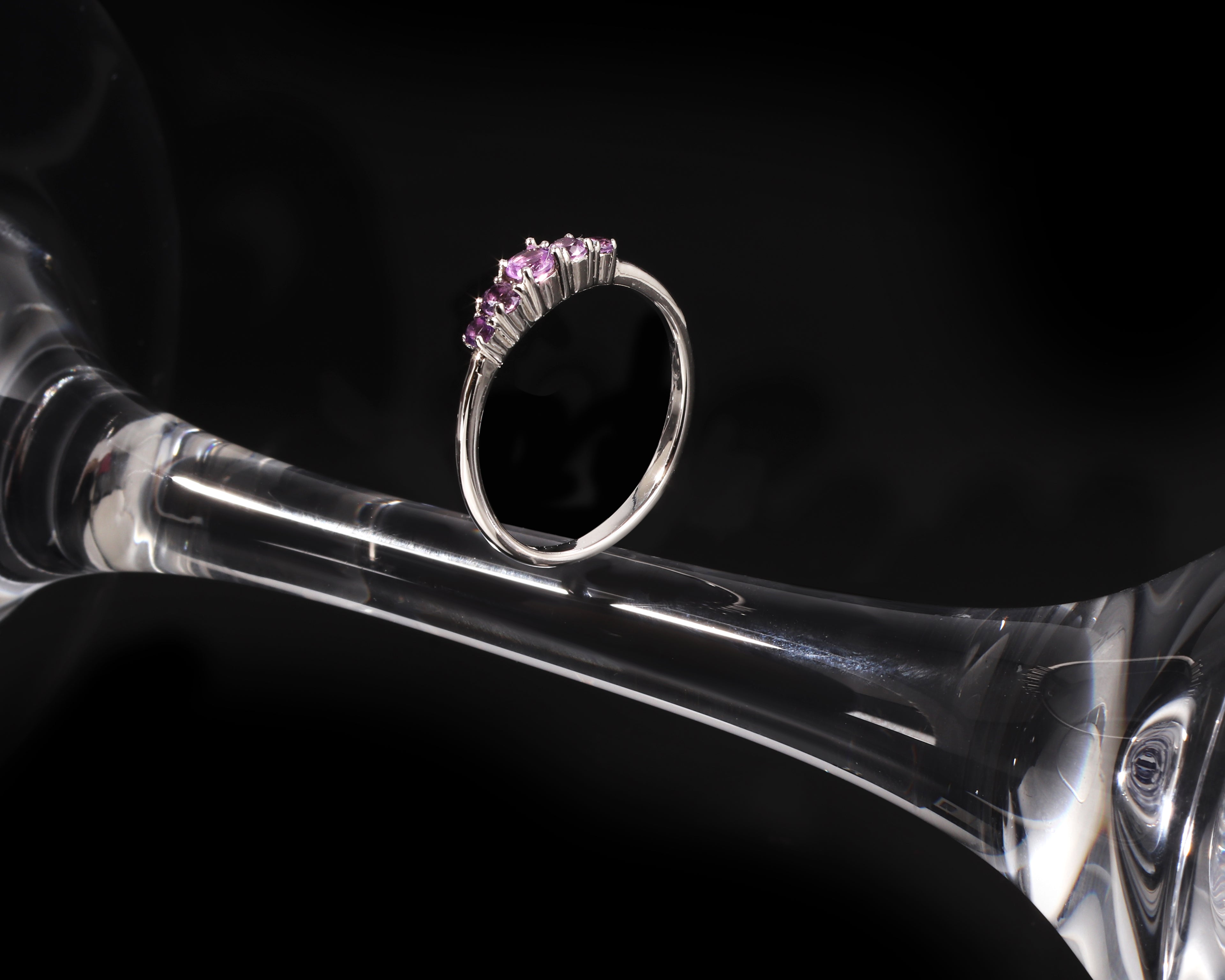 A silver ring with a row of small pink gemstones, displayed on the stem of a clear glass goblet against a black background.