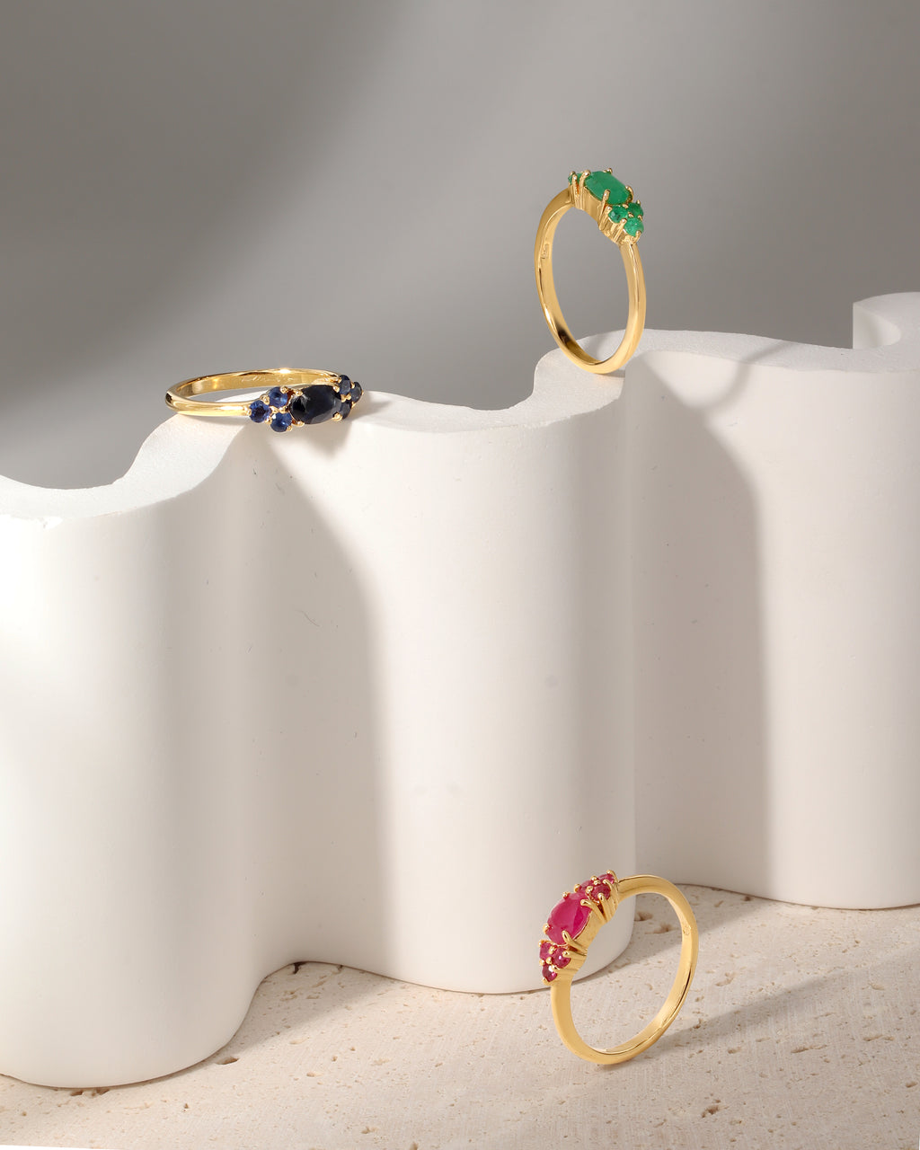 Three gold rings displayed on a sculptural white pedestal against a soft gray background. One ring has a green stone, one has blue and white stones, and one has a pink stone.