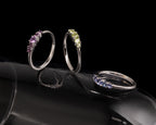 Three silver rings with colored gemstones displayed on a glossy black surface against a black background. 