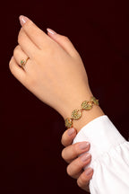 A close-up of a person's hand and wrist wearing a gold link bracelet with oval textured links, alongside a matching gold ring on the finger, set against a dark burgundy background.