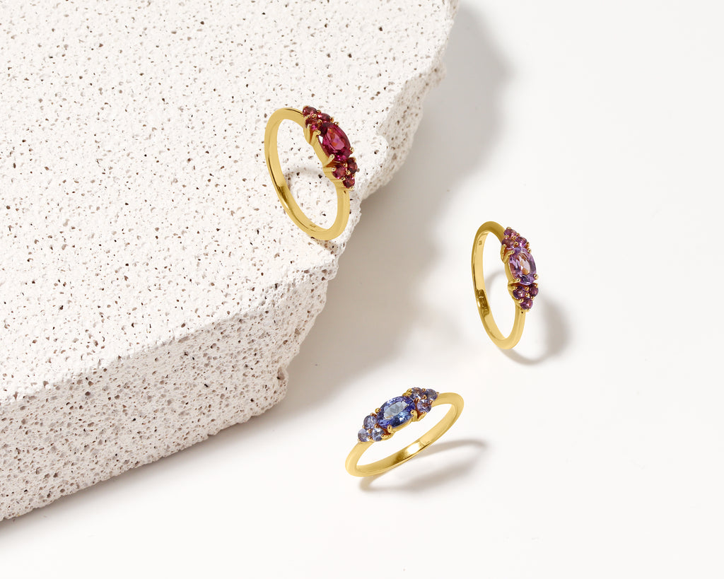 Three gold rings with oval-cut gemstones in red, purple, and blue, arranged on a white surface next to a textured white stone slab.