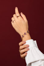 A close-up of a person's hands and wrist wearing jewelry against a dark burgundy background. 