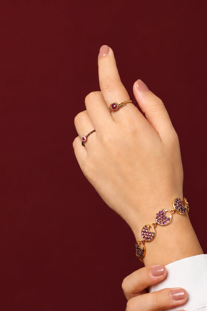 A close-up of a person's hands and wrist wearing jewelry against a dark burgundy background. 