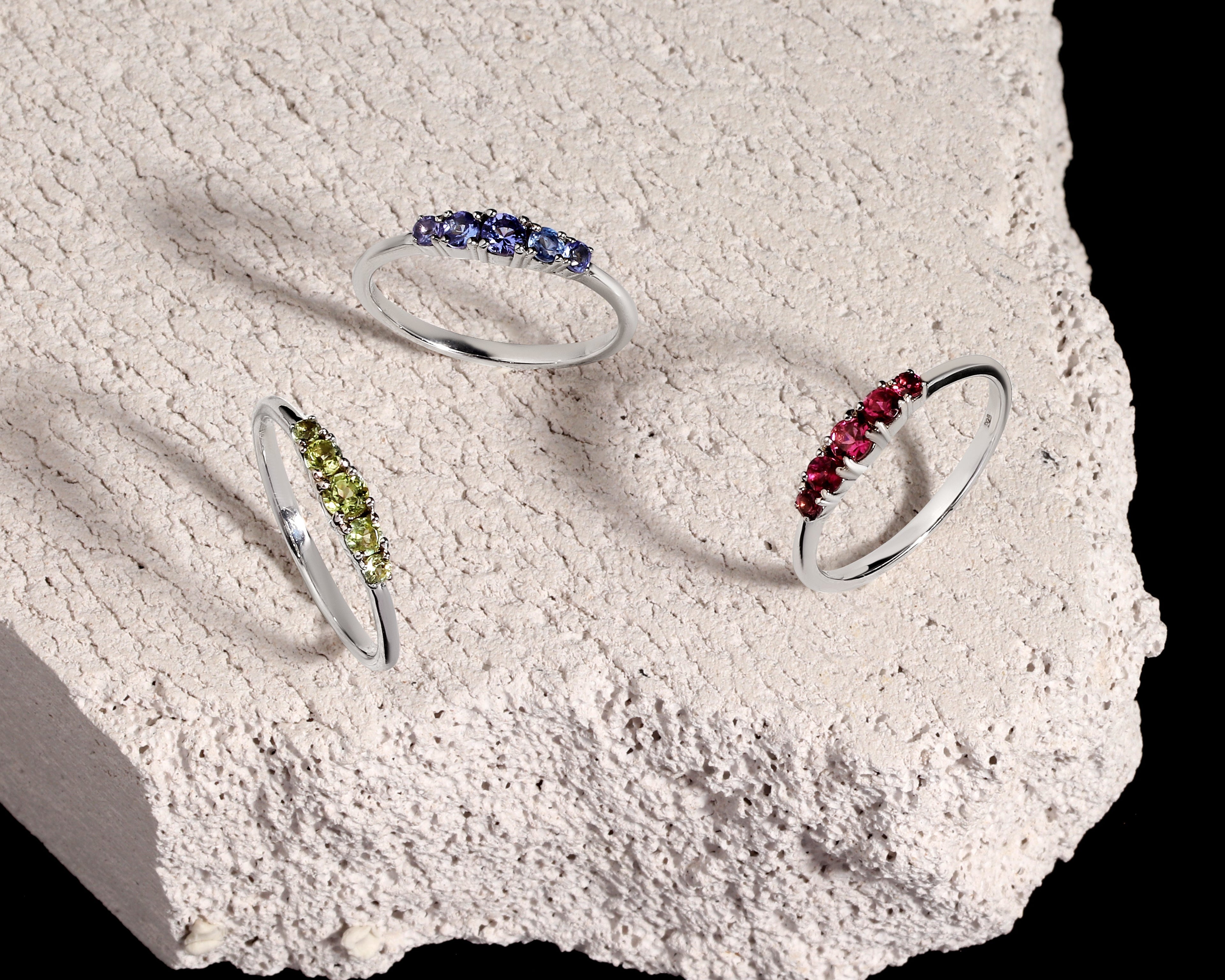 Three silver rings with small colored gemstone clusters are displayed on a rough white stone slab against a black background. One ring has blue stones, one has green stones, and one has red stones.