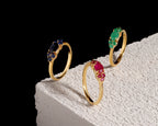 Three gold rings with marquise-cut gemstones in blue, green, and pink, displayed on a textured white block against a black background.
