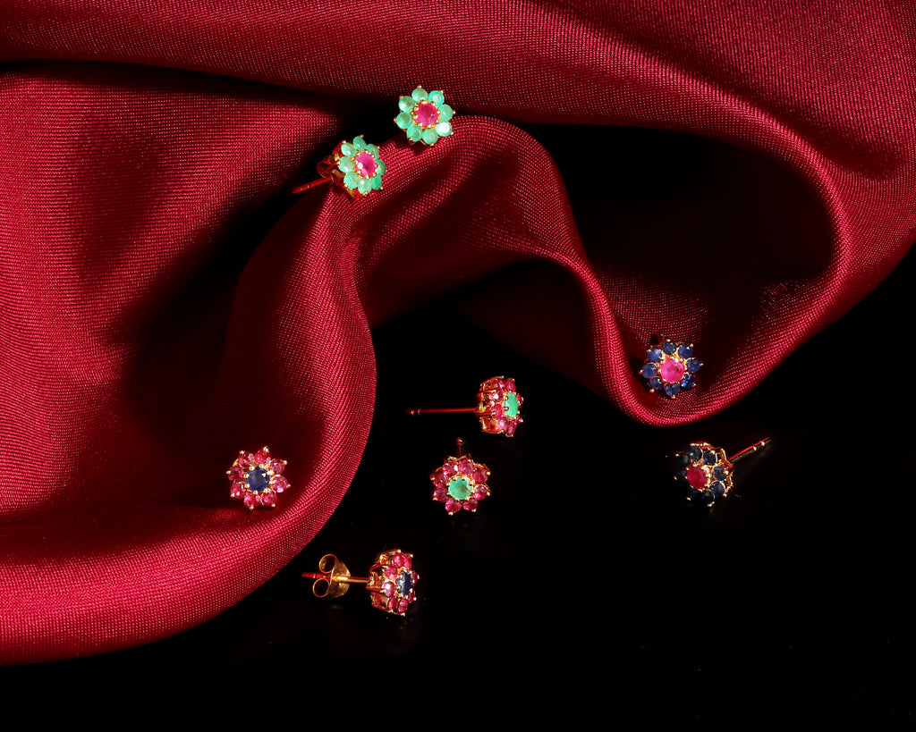A set of colorful flower-shaped stud earrings arranged on draped dark red satin fabric against a black background.