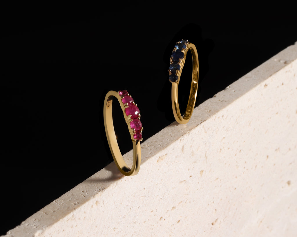 Two slim gold rings leaning against a textured white surface on a black background. One ring is set with bright pink gemstones, and the other is set with dark blue gemstones.