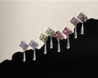 A set of seven silver teardrop dangle earrings with floral four-stone clusters in purple, green, and pink, arranged diagonally against a split beige and black background.