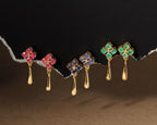 Four pairs of gold dangle earrings with floral tops in pink, blue, and green stones, displayed against a dark torn-paper background.