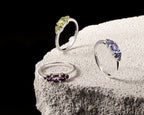 Three silver rings with gemstone clusters are displayed on a textured light gray stone pedestal against a black background. 