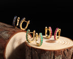 Four gold hoop earrings with colored gemstone borders in blue, green, pink, and gold, displayed on a rustic wood slice against a black background.