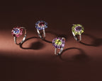 Four silver rings with halo gemstone designs are displayed on a dark brown background. 