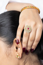 A close-up lifestyle shot of a person wearing a gold ring with a central red stone and a halo of small green and red stones, along with a matching gold bangle and earring.