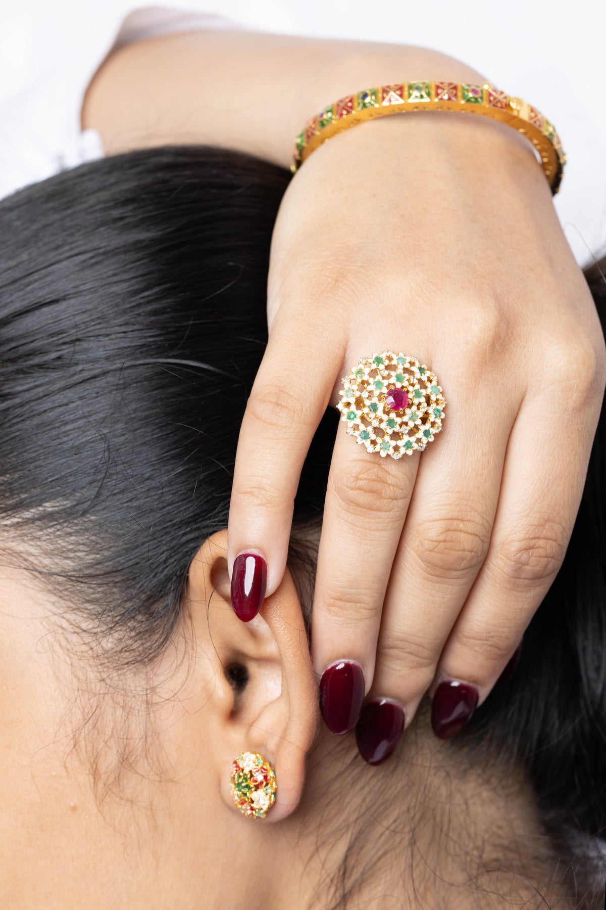 A close-up lifestyle shot of a person wearing a gold ring with a central red stone and a halo of small green and red stones, along with a matching gold bangle and earring.