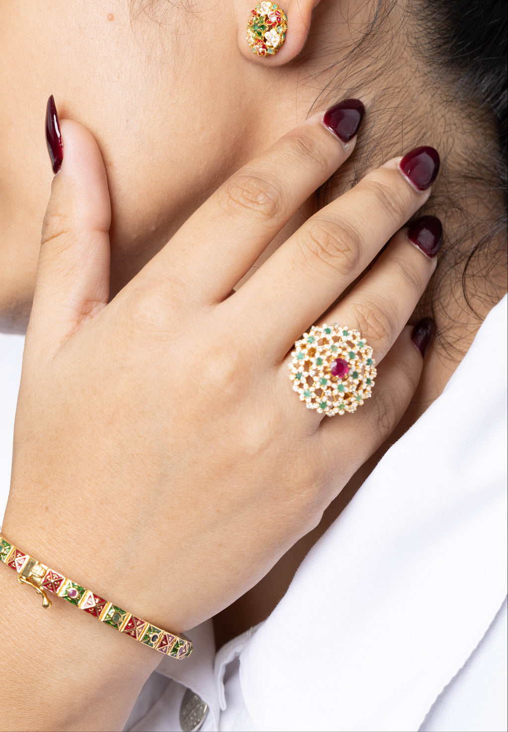 A close-up lifestyle shot of a woman wearing a gold-plated Kundan jewelry set, including a ring with a central red stone, matching stud earring, and bracelet, with her hand resting near her neck.