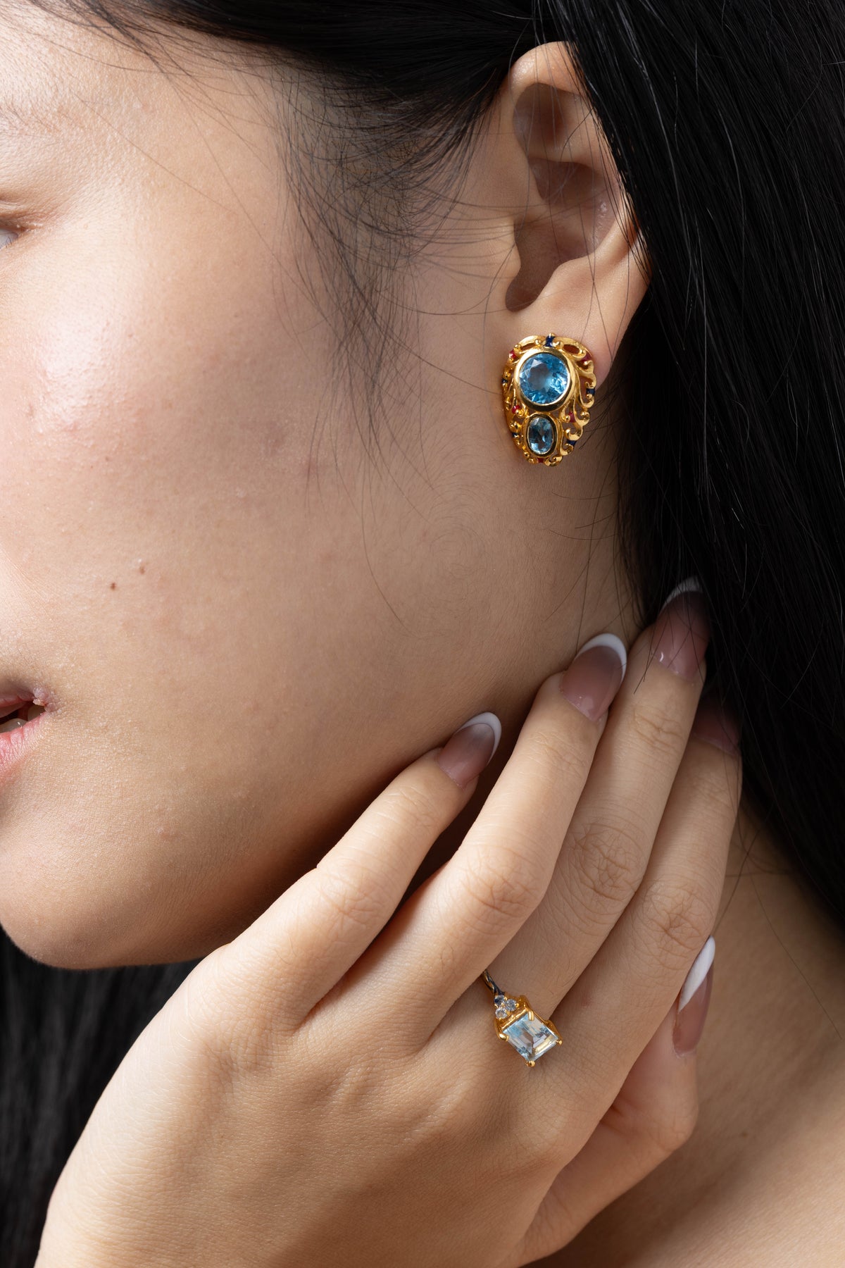 A close-up lifestyle shot of a woman wearing a gold-toned stud earring with blue oval and round stones, with her hand resting near her neck.