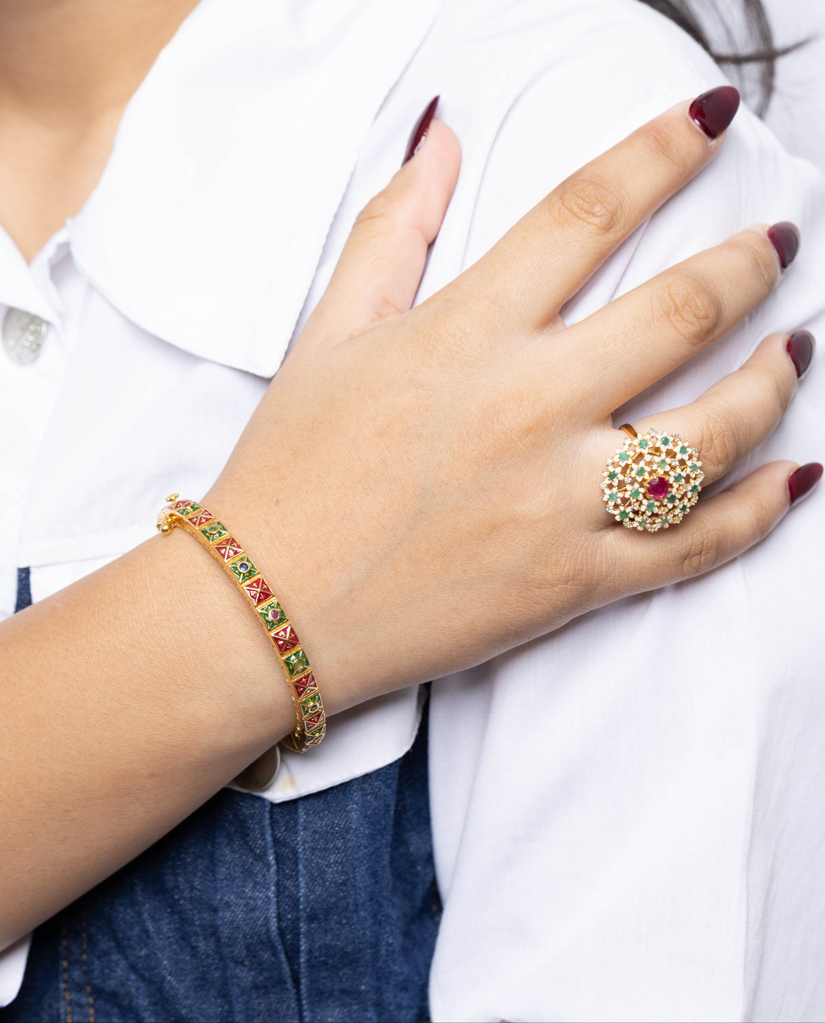 A close-up lifestyle shot of a person wearing a gold-plated bracelet with red and green stones on their wrist and a matching gold ring on their finger, paired with a white collared shirt and denim skirt.