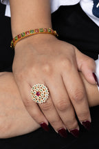 A close-up of a person's hands wearing jewelry, featuring a large gold ring with a white and red floral mandala design on the ring finger, and a gold bangle with red and green patterned stones on the wrist.