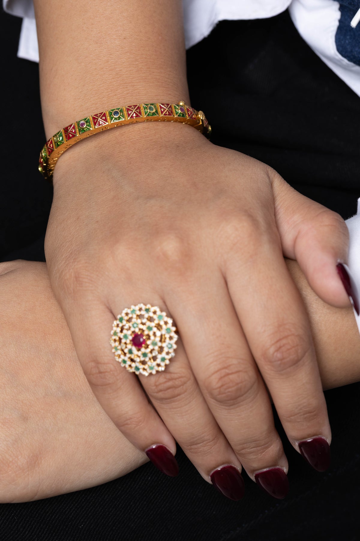 A close-up of a person's hands wearing jewelry, featuring a large gold ring with a white and red floral mandala design on the ring finger, and a gold bangle with red and green patterned stones on the wrist.