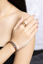 A close-up lifestyle shot of a woman's hand and shoulder wearing a gold ring and a gold bangle bracelet, both decorated with colorful enamel and small stones.
