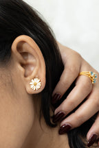 A close-up of a model's ear wearing a small daisy-shaped stud earring, with her hand resting near her head and a gold ring with blue stones visible on one finger.