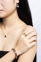 A close-up lifestyle shot of a model wearing a gold jewelry set featuring a necklace, matching drop earring, ring, and bracelet, all decorated with colorful enamel details.