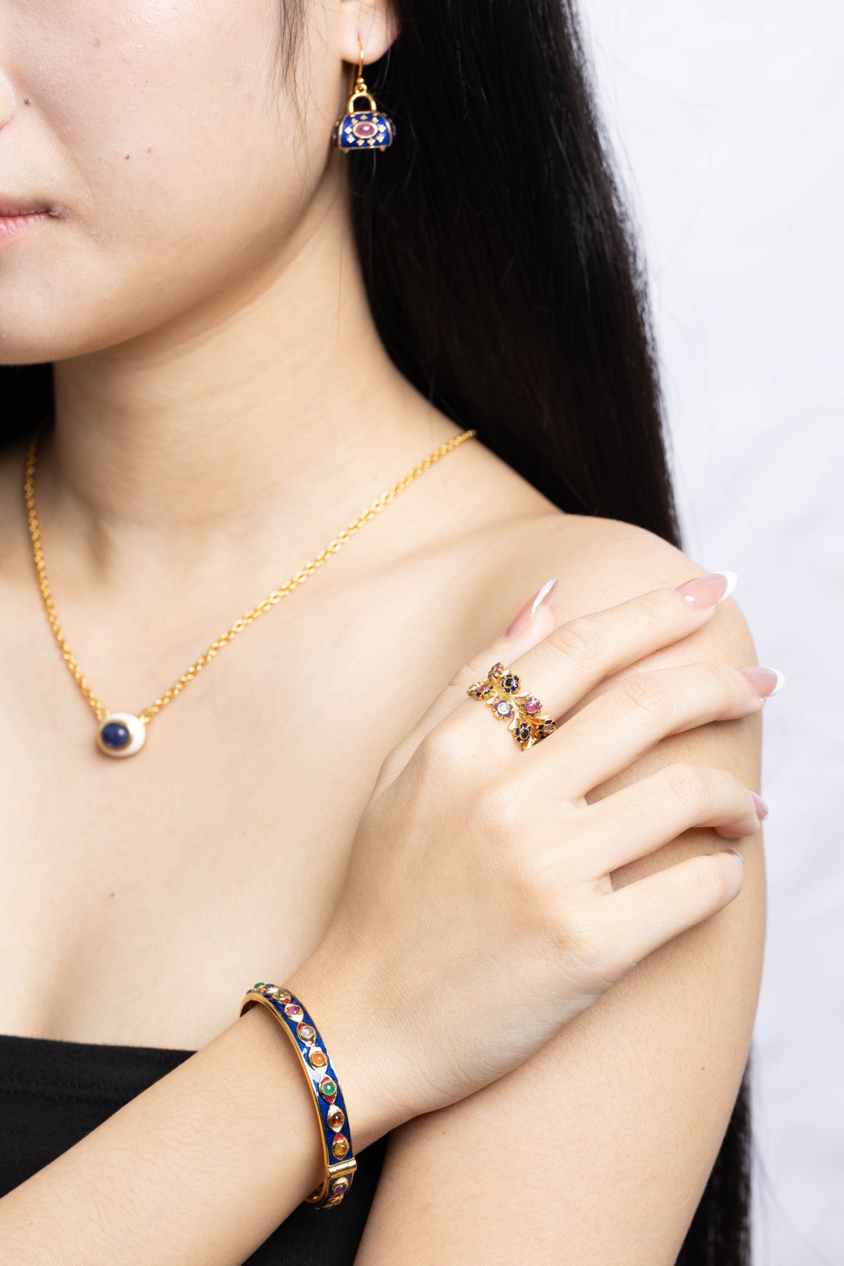 A close-up lifestyle shot of a model wearing a gold jewelry set featuring a necklace, matching drop earring, ring, and bracelet, all decorated with colorful enamel details.
