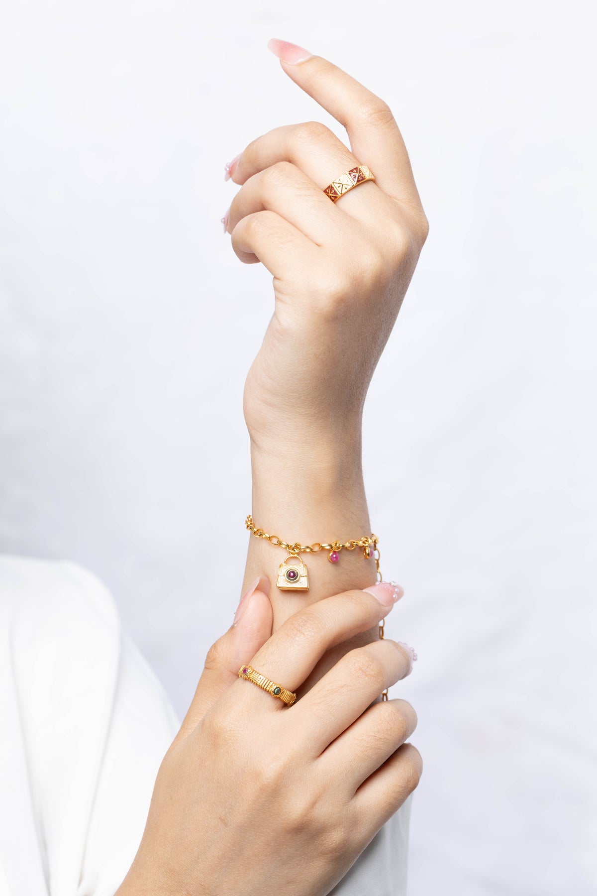 A close-up of a person's hands and wrist wearing gold jewelry, including a gold chain bracelet with a small padlock charm, a gold ring, and a gold signet-style ring, against a soft white background.