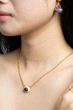 Close-up of a woman wearing a delicate gold chain necklace with a circular pendant featuring a deep blue center and a gold border, along with a matching dangling earring.