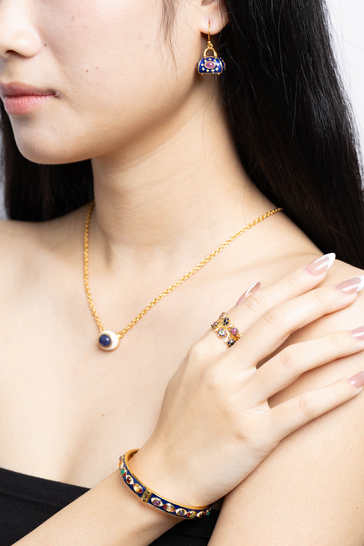 A close-up lifestyle shot of a model wearing a gold jewelry set, including a delicate gold chain necklace with a round blue stone pendant, a matching gold ring, and a gold bracelet with blue and red enamel details.