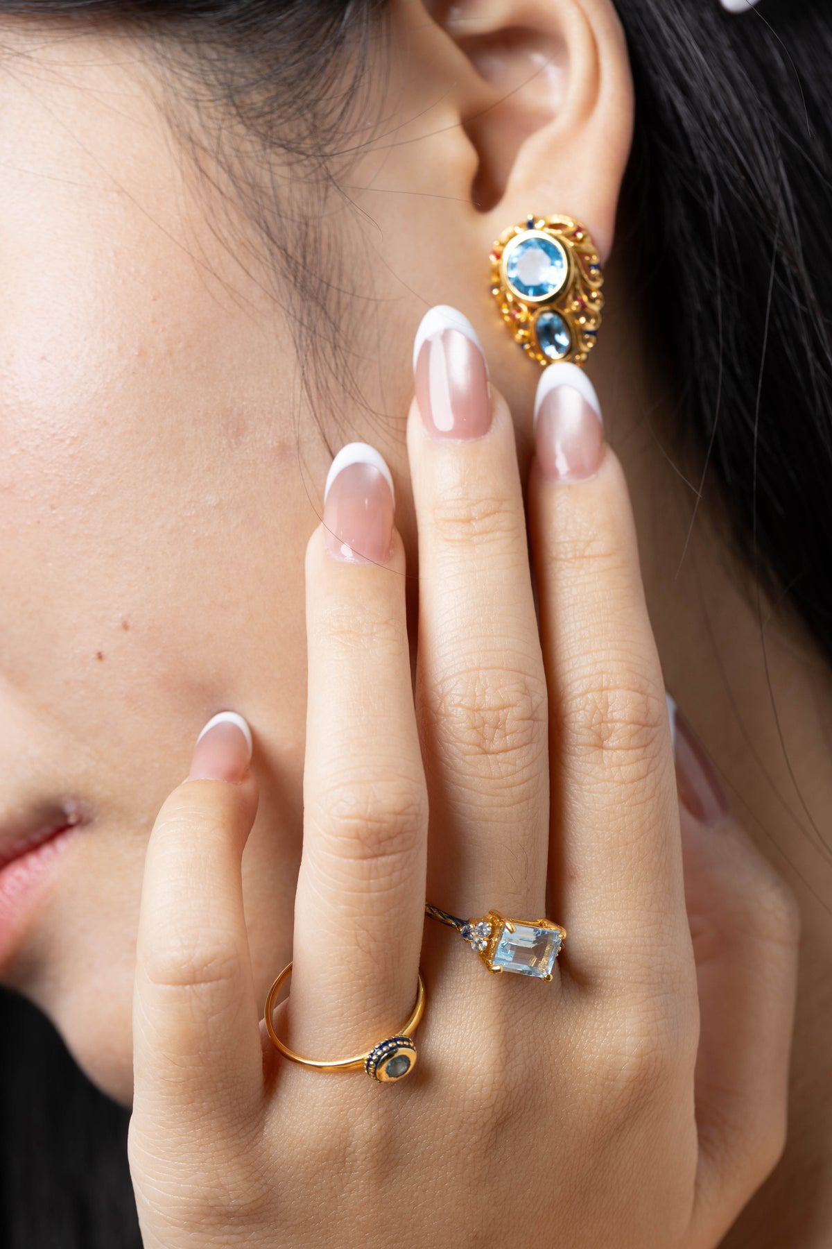 Close-up of a person's hand and ear wearing multiple gold rings and an earring with light blue gemstones.
