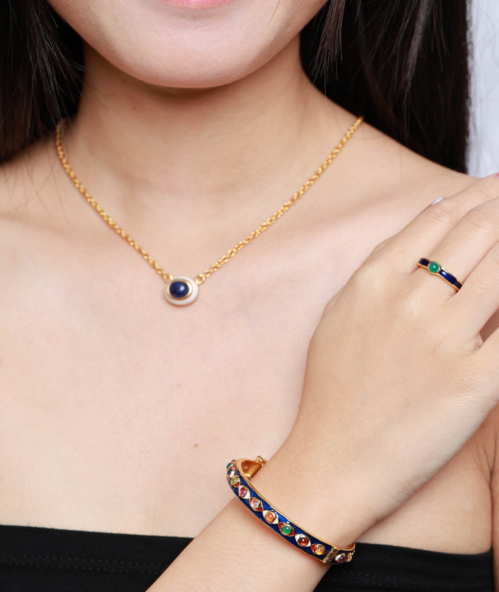 A close-up of a model wearing a gold jewelry set, featuring a delicate gold chain necklace with a circular navy blue and white evil eye pendant, a matching gold ring with colorful gemstone accents, and a gold bangle bracelet with a dark inlay and colorful stones.