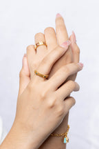 A close-up of two hands clasped together, showcasing gold jewelry including a gold chain bracelet with a small light blue pendant and gold rings on the fingers, set against a clean white background.