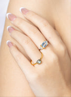 A close-up of a woman's hand with a French manicure, wearing two gold rings. One ring features a rectangular light blue gemstone, and the other is a delicate gold band with a small round clear stone.