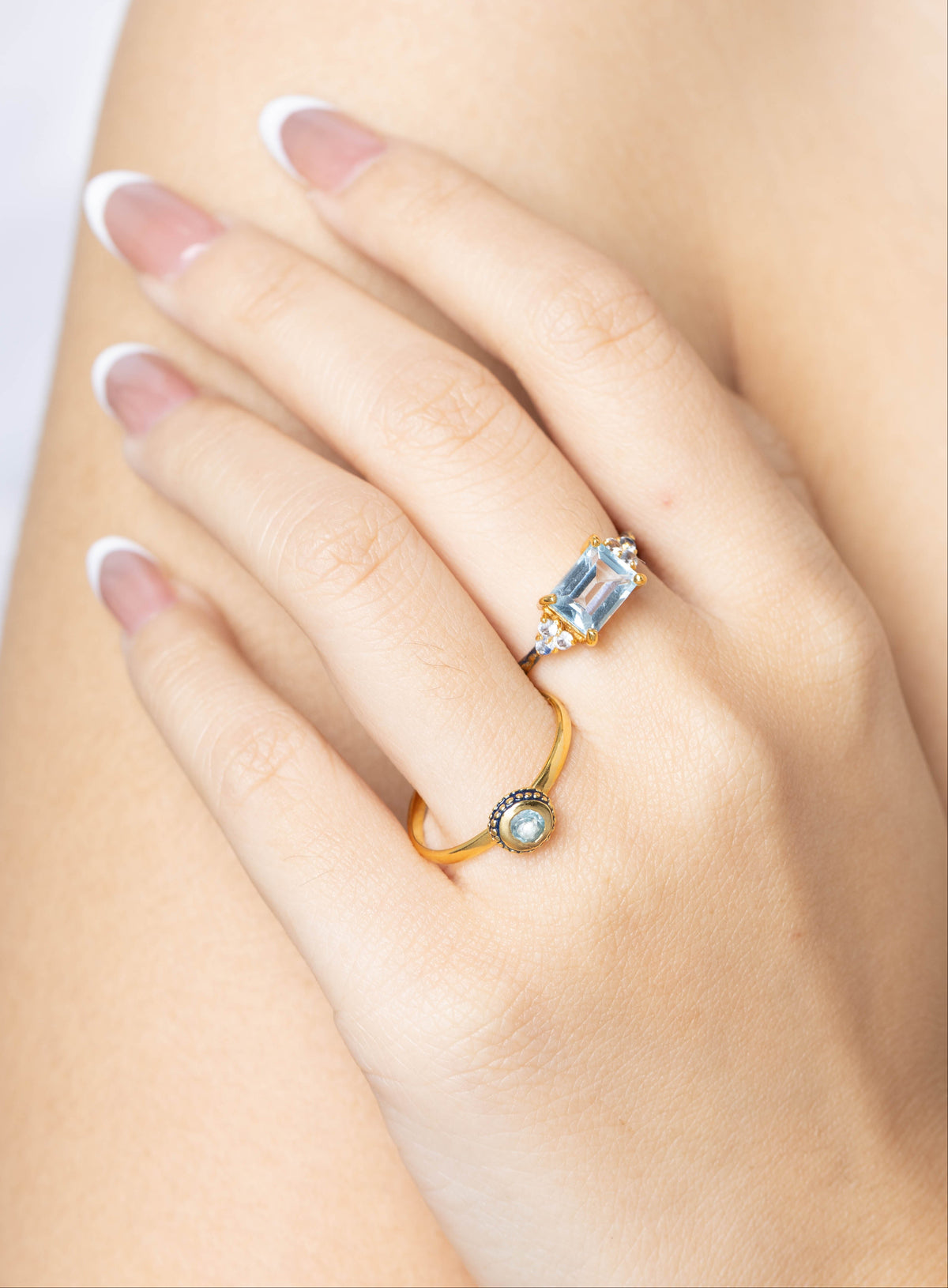 A close-up of a woman's hand with a French manicure, wearing two gold rings. One ring features a rectangular light blue gemstone, and the other is a delicate gold band with a small round clear stone.