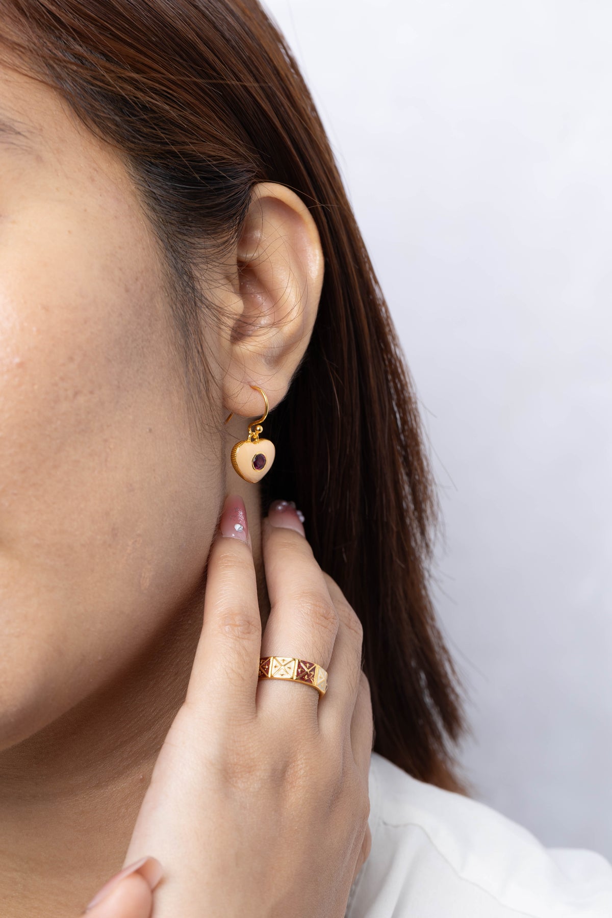 A close-up of a woman wearing a small gold heart-shaped drop earring with a dark center, along with a gold ring on her finger, against a light gray background.