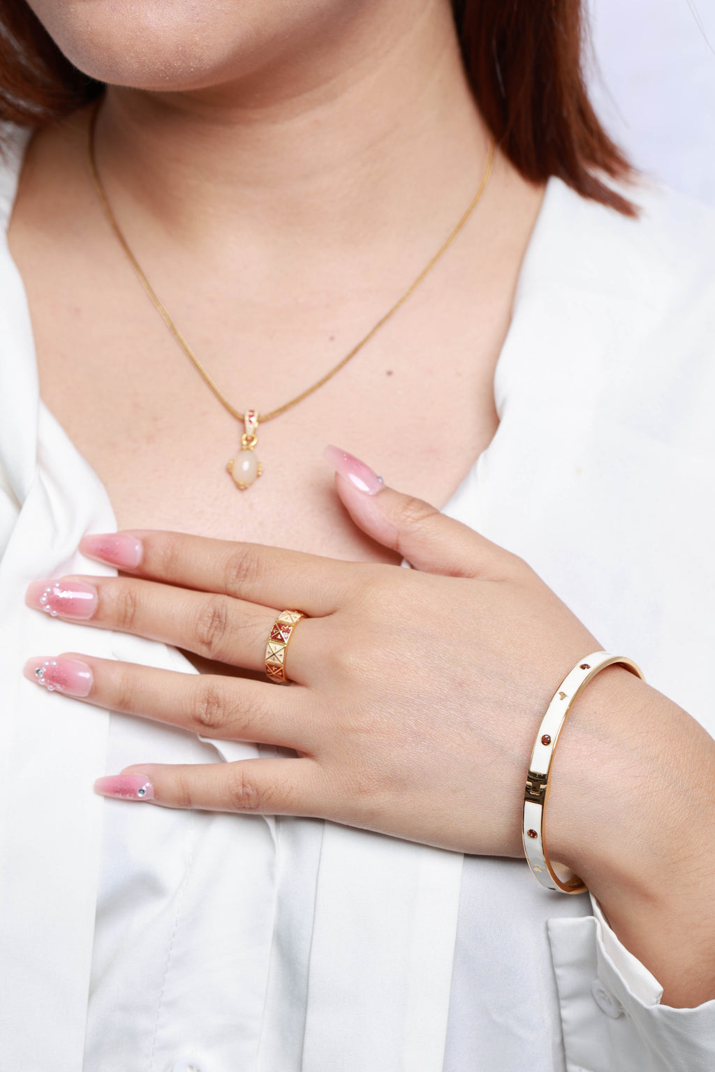 A close-up of a woman wearing a delicate gold necklace with a pearl-like pendant, a gold ring, and a white bangle with gold dots, styled with a white blouse.