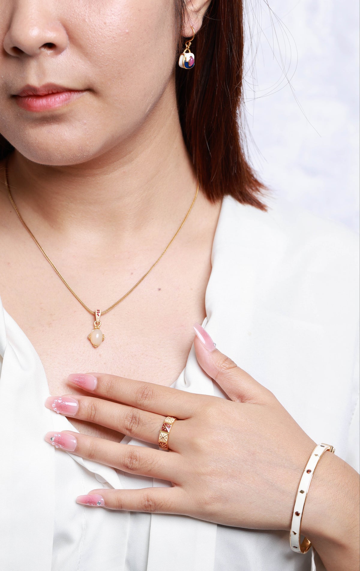 A close-up lifestyle shot of a woman wearing a delicate gold necklace with a small heart-shaped pendant, a gold ring, and a white beaded bracelet, wearing a white button-down shirt.