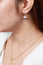 A close-up lifestyle shot of a woman wearing a delicate gold dangle earring with a colorful enamel face and a matching gold necklace.
