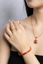 A close-up lifestyle shot of a model wearing a gold necklace with a red and green turtle pendant, a matching red and gold bracelet, and a gold ring.