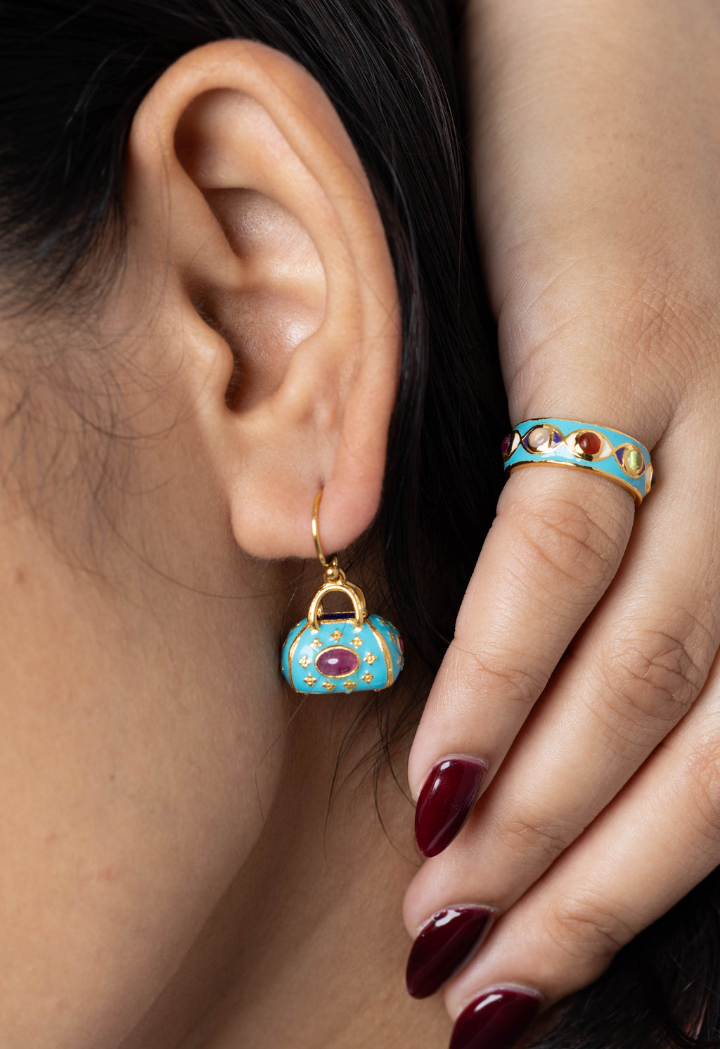 Close-up of a model wearing a small turquoise and gold handbag-shaped drop earring and a matching turquoise and gold ring with red gemstone accents.
