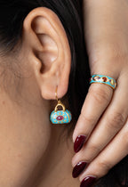 Close-up of a model wearing a small turquoise and gold handbag-shaped drop earring and a matching turquoise and gold ring with red gemstone accents.