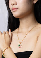 A woman wearing a gold necklace with a small floral pendant and a matching gold bracelet, styled with a black strapless top against a light gray background.