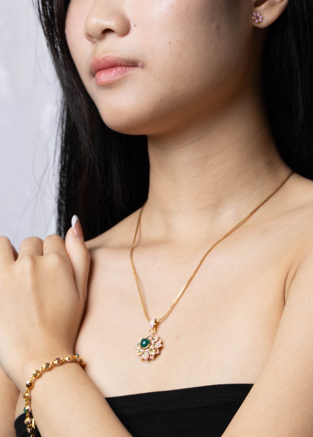 A woman wearing a gold necklace with a small floral pendant and a matching gold bracelet, styled with a black strapless top against a light gray background.