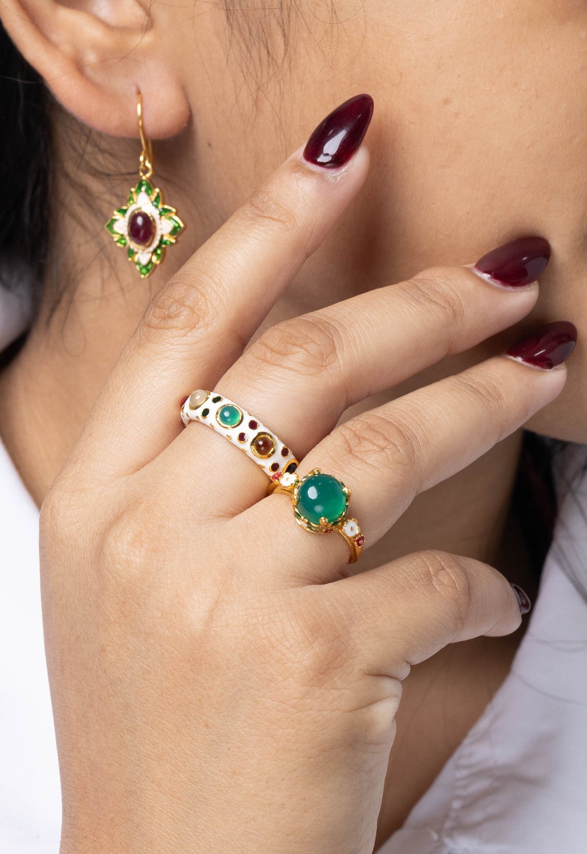 A close-up lifestyle shot of a woman wearing gold jewelry, featuring a gold ring with green and white stones on her finger and a matching gold dangle earring with a red stone on her earlobe.