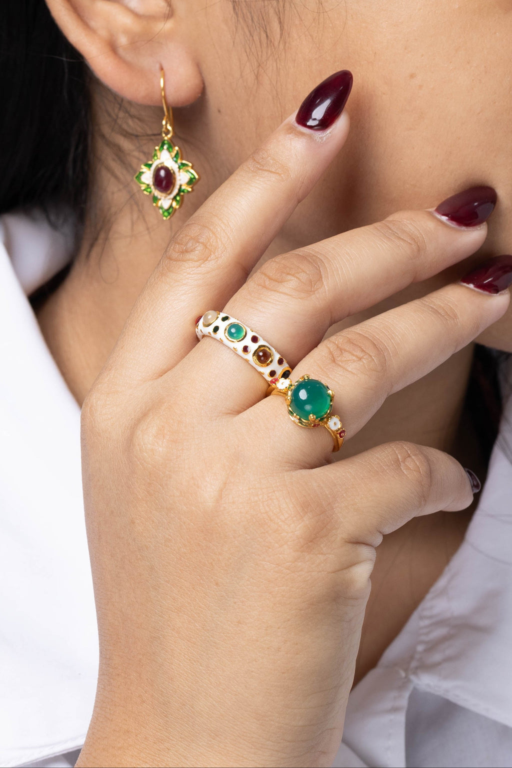 A close-up lifestyle shot of a woman wearing gold jewelry, featuring a gold ring with green and white stones on her finger and a matching gold dangle earring with a red stone on her earlobe.