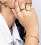 A woman in a white collared shirt shown in profile, wearing gold rings, a gold bracelet, and green earrings, against a plain light gray background.
