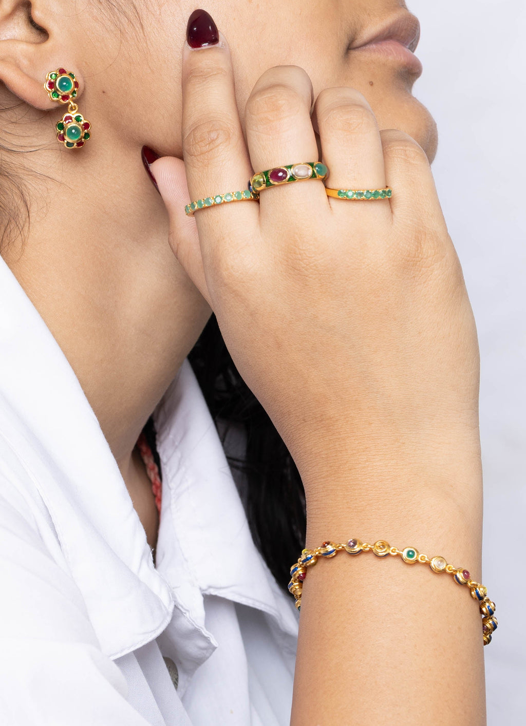 A woman in a white collared shirt shown in profile, wearing gold rings with green stones, a matching gold bracelet, and a small gold earring with green stones, against a light gray textured background.