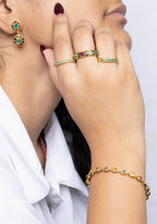 A woman in a white collared shirt shown in profile, wearing gold rings with green stones, a matching gold bracelet, and a small gold earring with green stones, against a light gray textured background.