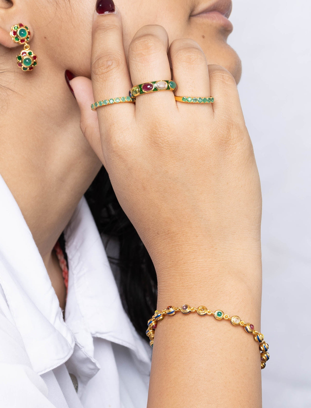 A woman in a white collared shirt shown in profile, wearing gold rings on her fingers and a gold bracelet on her wrist, with a small green and gold earring visible on her ear. 